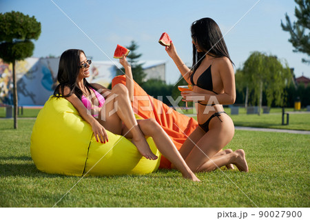 Side view of two pretty girl sitting on armchair bags, relaxing. Seductive young females smiling, talking, drinking cocktails, eating watermelon. Concept of summertime and youth. 90027900