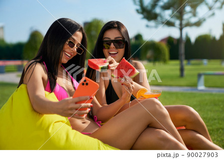 Front view of two pretty brunette women sitting on armchair bags, taking selfie. Seductive young women relaxing, smiling, talking, eating watermelon. Concept of summertime. 90027903