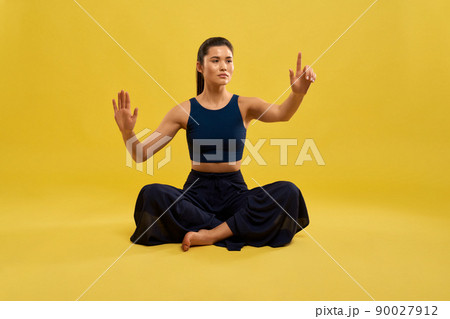 Front view of sporty girl sitting with crossed legs on floor, raising hands, holding finger up, looking forward. Young female practicing yoga pose. Isolated on yellow background. Front view of sporty girl sitting with crossed legs on floor, raising hands, holding finger up, looking forward. Young female practicing yoga pose. Isolated on yellow background. 90027912