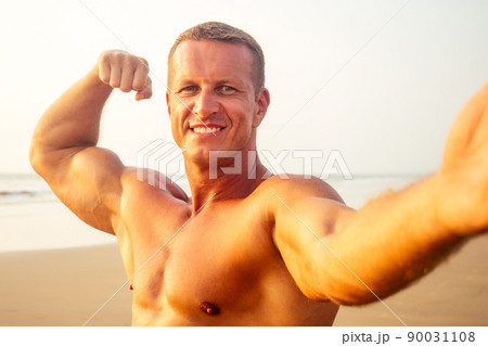 handsome successful male athlete fitness model photographing selfie portrait on phone beach paradise sunset. man on vacation in the tropic talking on the smartphone online with friends and family 90031108