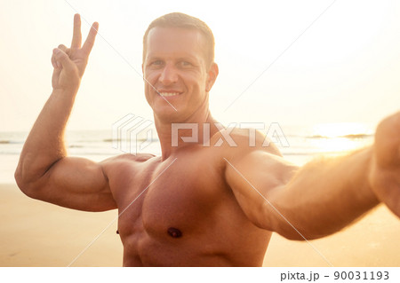 handsome successful male athlete fitness model photographing selfie portrait on phone beach paradise sunset. man on vacation in the tropic talking on the smartphone online with friends and family 90031193