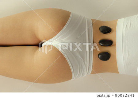 Cropped image of female body in underwear with warm stones on it isolated over grey studio background. Stone massage treatment 90032841