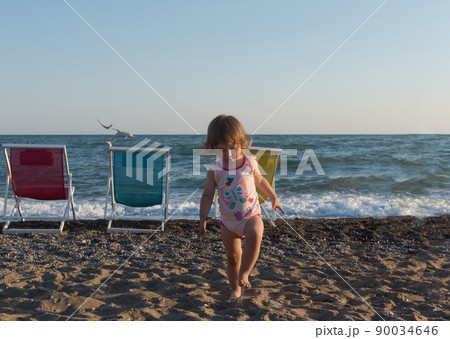 Little girl in sitting on sand and playing with bucket and spade at sea beach in warm sunny summer day. Closeup. Side view. Little girl in sitting on sand and playing with bucket and spade at sea beach in warm sunny summer day. Closeup. Side view. 90034646
