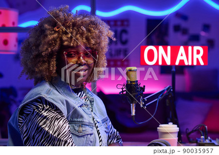 Horizontal medium close-up of joyful young African American woman wearing fashionable outfit sitting in broadcasting studio smiling at camera 90035957