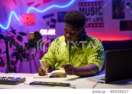 Portrait of creative young gen Z African American man sitting at desk in home studio working on broadcast or podcast text 90036027