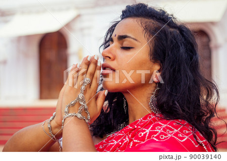 Young Indian woman in traditional sari red dress praying in a hindu temple goa india Hinduism.girl performing namaste gesture catholicism Delhi Street holi festival.om yoga meditation female model 90039140