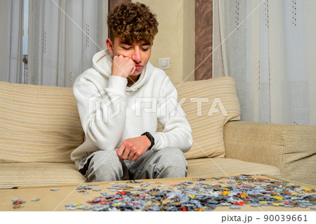 Young man with curly hair sitting on the sofa at home trying to assemble a puzzle with many pieces Young man with curly hair sitting on the sofa at home trying to assemble a puzzle with many pieces 90039661