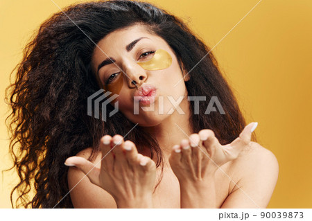 Happy enjoyed pretty Latin female with gold hydrogel patches under eye blowing kiss posing isolated over yellow background. Cosmetic product ad Natural beauty concept Studio portrait Copy space Happy enjoyed pretty Latin female with gold hydrogel patches under eye blowing kiss posing isolated over yellow background. Cosmetic product ad Natural beauty concept Studio portrait Copy space 90039873