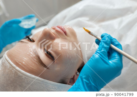Beautiful brunette woman enjoying applying cosmetic mask with closed eyes in sunny spa center. Relaxing treatment and medicine concept. 90041799