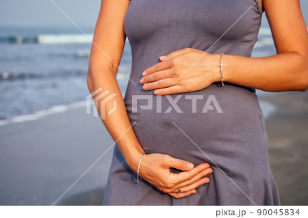beautiful pregnant woman in a blue grey tight dress is covered with hat on the beach touching her belly with love and care on the background sea. 90045834