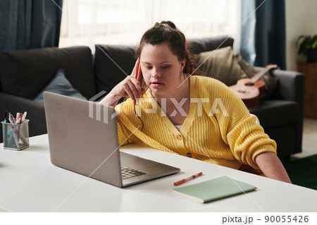 Young Woman with Down syndrome sitting relaxed at table at home with laptop on it talking on phone with friend or co-worker 90055426
