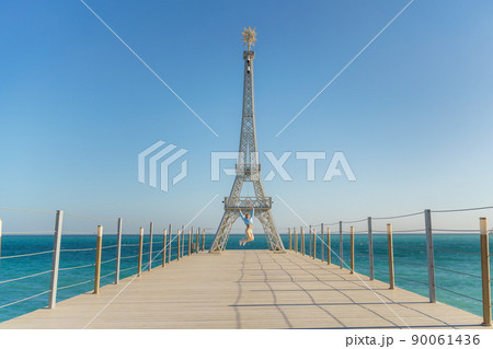 Large model of the Eiffel Tower on the beach. A woman walks along the pier towards the tower, wearing a blue jacket and white jeans. 90061436