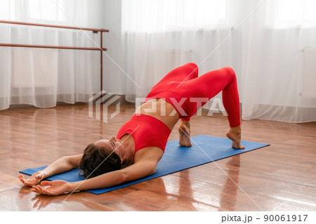 A young woman fitness instructor in red Sportswear Leggings and Top stretching in the gym before her pilates, on a yoga mat near the large window on a sunny day, female fitness yoga routine concept. 90061917