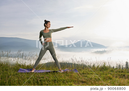 Fit young woman in sport clothes practicing yoga during morning time among green nature. Amazing view of summer mountains on background. 90068800
