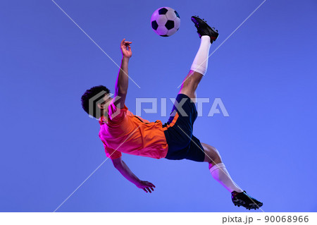 Studio shot of male professional football, soccer player practicing with ball isolated on purple background. Concept of sport, match, active lifestyle, goal and hobby Studio shot of male professional football, soccer player practicing with ball isolated on purple background. Concept of sport, match, active lifestyle, goal and hobby 90068966