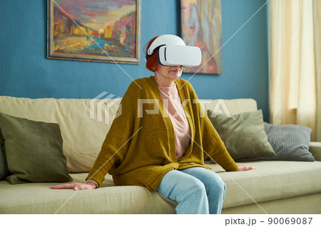 Mature woman in vr goggles sitting on sofa in living room and watching virtual reality 90069087
