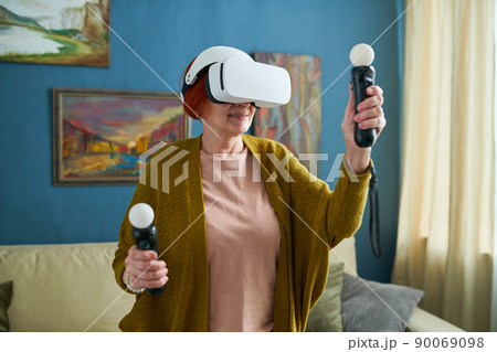 Mature woman in goggles holding joysticks in her hands and gesturing during virtual reality game at home 90069098