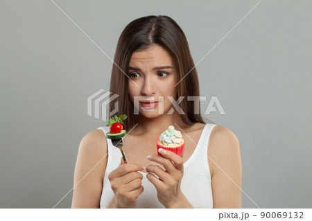 Sad woman iholding vegetable and sweets on grey background 90069132