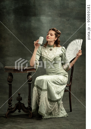 Vintage portrait of young beautiful girl in gray dress of medieval style drinking tea isolated on dark background. Comparison of eras concept, flemish style. Art, beauty 90069181