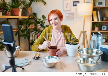 Senior woman making her blog about gardening, she sitting at table and transplanting flowers in other pots n camera of smartphone 90069211