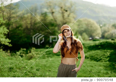 A beautiful woman in sportswear and a cap walks against a green natural landscape and smiles in the sunlight 90070548