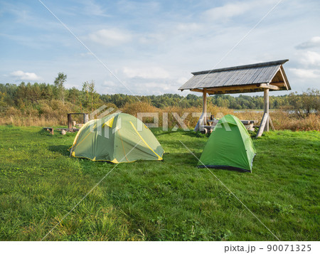 Two green touristic tents stand in camp. Wooden table, benches and fire pit for camping. Hiking in Russia. 90071325