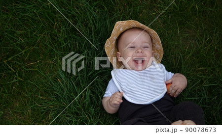 Small Happy Newborn child in summer panama hat Fall down laying on grass barefoot in Summer Sunny Day. Infant Kid Toddler Boy Smilling Face look at camera in Garden ouside Family Childhood Nature. 90078673