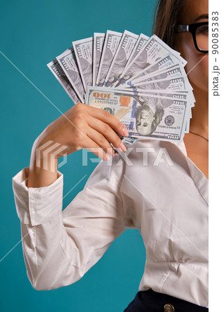 Brunette girl in glasses, wearing in a black short skirt and white blouse is posing holding a fan of hundred dollar bills against a blue background. 90085383