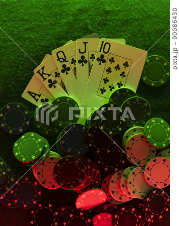 Close-up top view photo of royal flush and chips laying on green cover of playing table, against black background, under green and red neon lights. 90086430