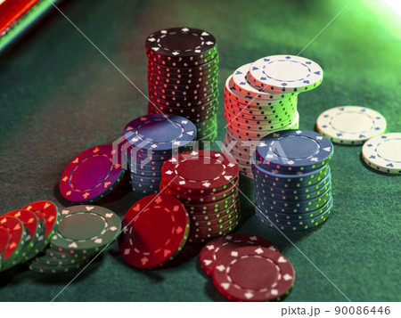 Close-up top view of colorful chips piles, some of them laying nearby on green cover of playing table, under green, red neon light. Black background. Close-up top view of colorful chips piles, some of them laying nearby on green cover of playing table, under green, red neon light. Black background. 90086446