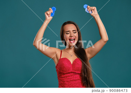 Brunette girl with a long hair, wearing a sexy red dress is holding a few gambling chips in her hands while posing against a blue background. 90086473