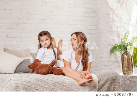 Indoor portrait of a beautiful mother with her charming little daughter posing against bedroom interior. Indoor portrait of a beautiful mother with her charming little daughter posing against bedroom interior. 90087797