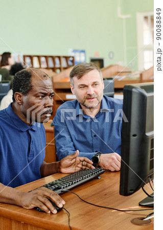 Professional teacher working with immigrants in international school sitting at desk next to mature Black man helping him to understand task on computer Professional teacher working with immigrants in international school sitting at desk next to mature Black man helping him to understand task on computer 90088849