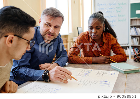 Mature English language teacher sitting at table with his students asking them questions during lesson 90088951