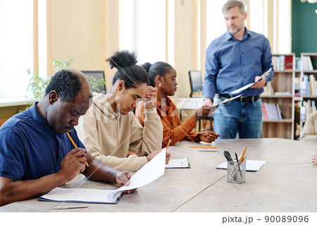 Group of modern Black immigrants attending classes sitting at table finishing doing English grammar test Group of modern Black immigrants attending classes sitting at table finishing doing English grammar test 90089096