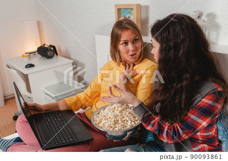 The concept of lesbian relationships. LGBT. Two young women are sitting on a bed, eating popcorn and using a laptop. The interior of the room in the background The concept of lesbian relationships. LGBT. Two young women are sitting on a bed, eating popcorn and using a laptop. The interior of the room in the background 90093861