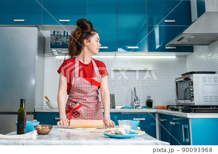 A young pretty housewife rolls out the dough with a rolling pin on the table in the kitchen and looks at the oven A young pretty housewife rolls out the dough with a rolling pin on the table in the kitchen and looks at the oven 90093968