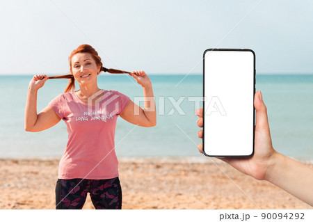 An adult woman in sportswear smiles and poses against the background of the sea, cheerfully spreading her hair with her hands. Mock up. Female hand holding a smartphone. Sports lifestyle concept 90094292