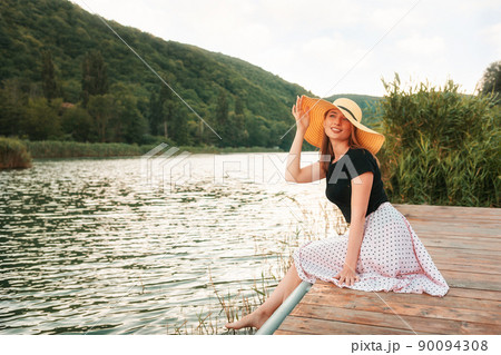 Summer vacation. A beautiful young woman poses elegantly, sitting on a pier by the sea and holding her hat. Landscape in the background Summer vacation. A beautiful young woman poses elegantly, sitting on a pier by the sea and holding her hat. Landscape in the background 90094308