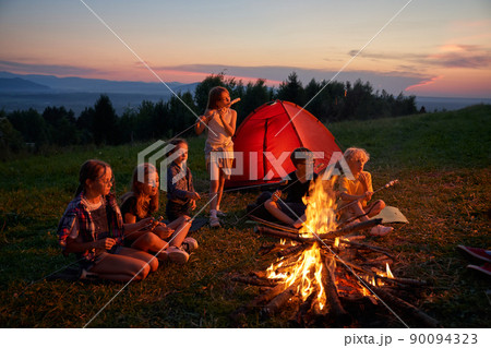 Side view of group of kids sitting eating in campsite in mountains. Six children having dinner, camping near fireplace with red tent on background. Concept of traveling. 90094323