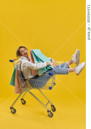 Side view of girl with curly hair doing shopping. Young female sitting in shopping cart, holding, hugging packages, looking at camera. Isolated on yellow studio background. 90094451