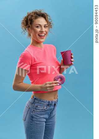 Side view of beautiful young female holding donaught and cup of tea or coffee. Pretty girl with curly hair standing, smiling, looking at camera. Isolated on blue background, Side view of beautiful young female holding donaught and cup of tea or coffee. Pretty girl with curly hair standing, smiling, looking at camera. Isolated on blue background, 90094453