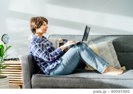 Smiled mixed race woman using laptop at home sitting on soft couch. Cozy workspace in modern interior with green plants. Remote work at home. Online education or entertainment. Copy space 90095900