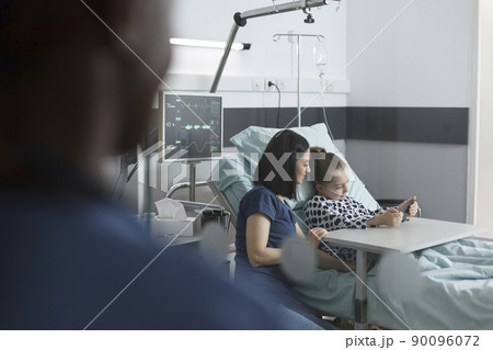 Mother sitting beside patient bed in pediatric clinic room while cute little girl playing on smartphone. Joyful young patient enjoying mobile phone entertainment while sitting in clinic recovery ward. 90096072