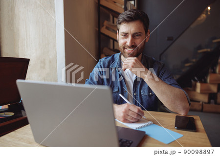 Portrait of Caucasian handsome happy man taking notes and looking at laptop in cafe. Freelancing. 90098897