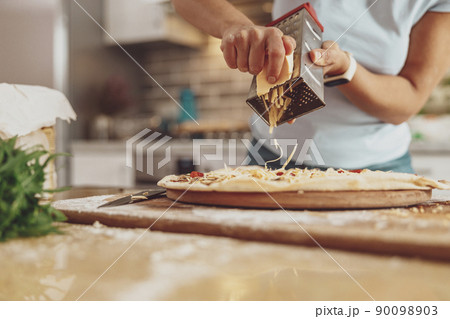 Women's hands grate cheese on a pizza lying on the table in the background of the kitchen 90098903