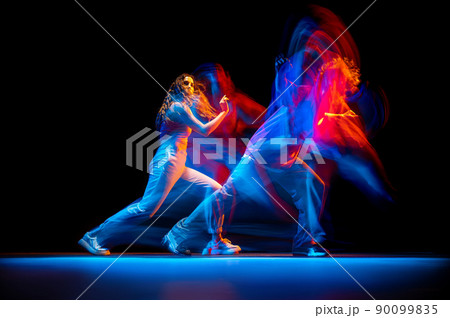 Dynamic portrait of stylish man and girl dancing hip-hop in sportive style clothes on dark background at dance hall in mixed neon light. Youth culture, breakdance, movement Dynamic portrait of stylish man and girl dancing hip-hop in sportive style clothes on dark background at dance hall in mixed neon light. Youth culture, breakdance, movement 90099835
