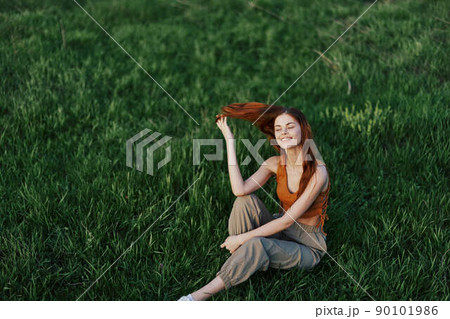 A woman enjoying the outdoors sitting in the park on the green grass in casual clothing with long flowing hair, lit by the bright summer sun without mosquitoes 90101986