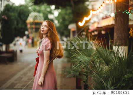 Young beautiful red-haired long-haired woman in a summer pink dress walks through the streets of a summer city. Young beautiful red-haired long-haired woman in a summer pink dress walks through the streets of a summer city. 90106352