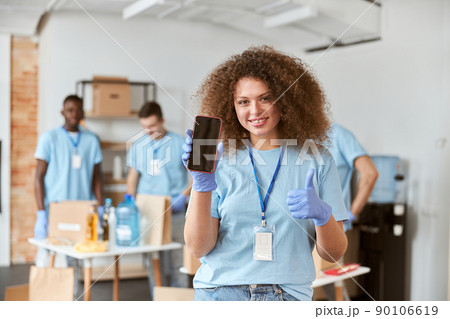 Young woman, volunteer in blue uniform holding, showing smartphone to camera and smiling while standing indoors. Team sorting, packing items in the background 90106619